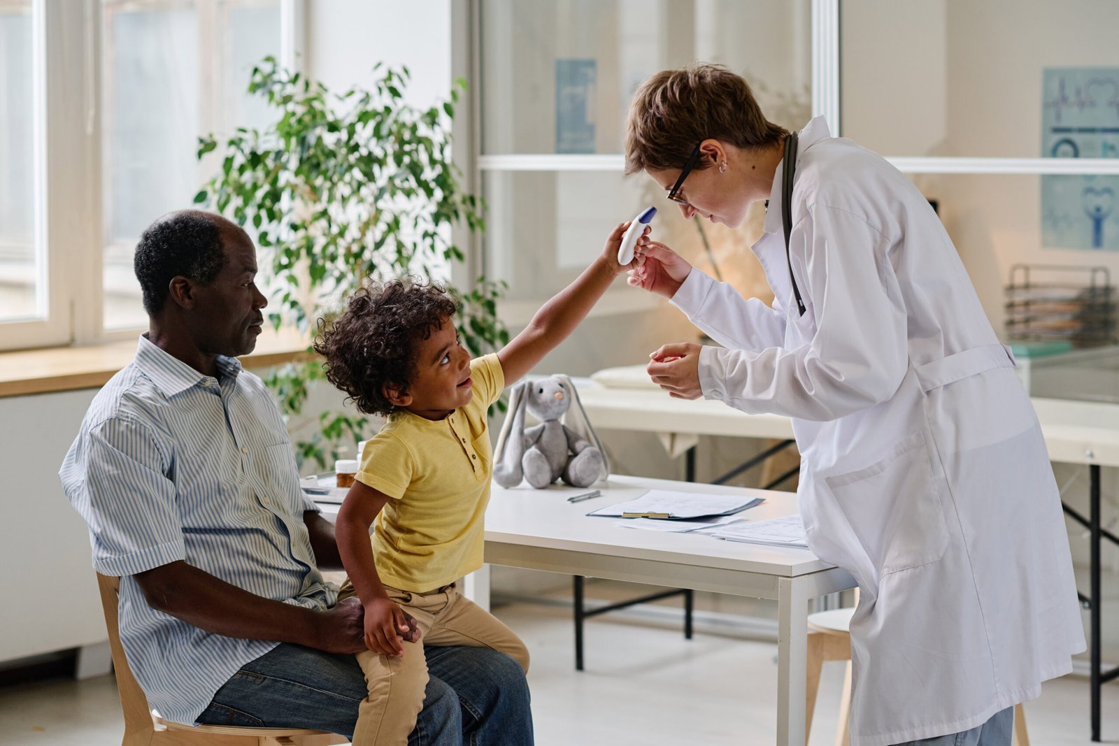 Female doctor trying to examine child with special medical equipment while he sitting with his dad at clinic
